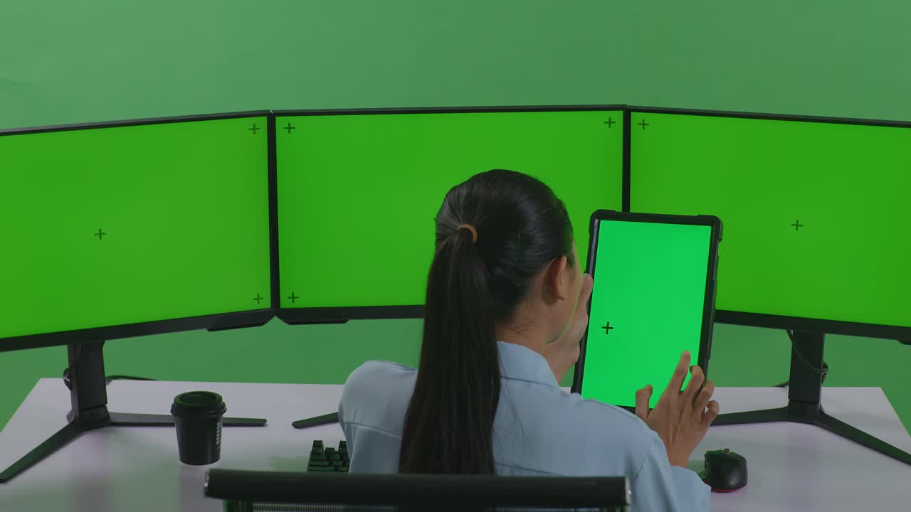 태블릿과 여러 컴퓨터 모니터로 작업하는 아시아 여성의 뒷면은 녹색 스크린 스튜디오에서 녹색 화면 모형을 가지고 있습니다.
