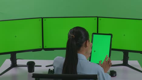 vista trasera de una mujer asiática trabajando con una tableta y varios monitores de computadora con una pantalla verde simulada en un estudio de pantalla verde