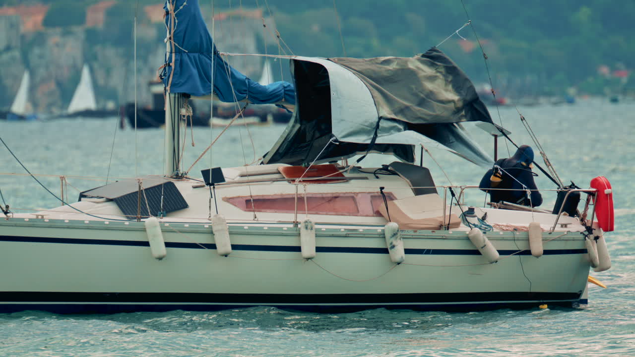 View of a small sailboat rocking on the waves with a lone sailor on deck, diver preparing to jump into sea