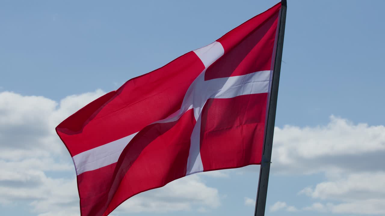 Rødt og hvidt korsflag, der dynamisk vajer udendørs i dagslys med blå himmel og skyer i baggrunden