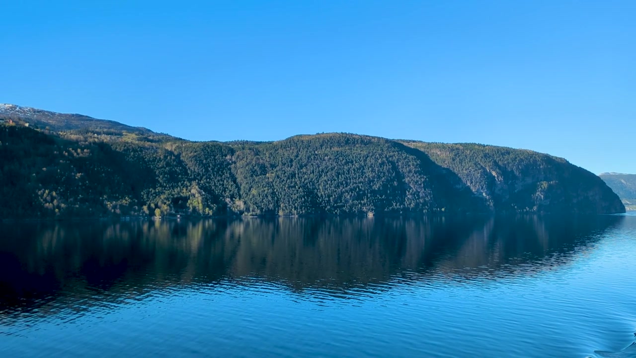 Spejlbilleder af skovklædt bjerg på roligt blåt vand i norsk fjord.