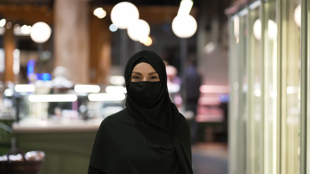 Ritratto di una donna con hijab e mascherina al supermercato, in piedi con il carrello della spesa.
