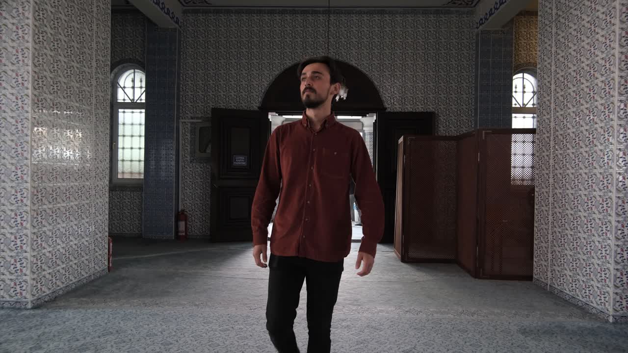 Vista frontal de um jovem barbudo caminhando pela porta da mesquita, pessoas visitam locais de culto históricos e culturais.