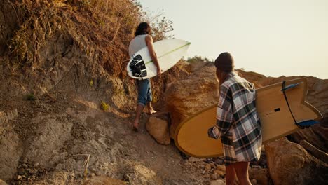 un chico moreno en una camiseta blanca junto con su novia en un sombrero verde y una camisa a cuadros caminan a lo largo de la costa rocosa a lo largo del mar y llevan tablas de surf con ellos en una mañana de otoño