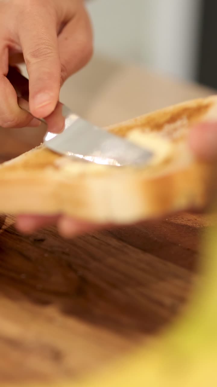 A hand spreads butter on a slice of bread using a knife, in a warmly lit kitchen