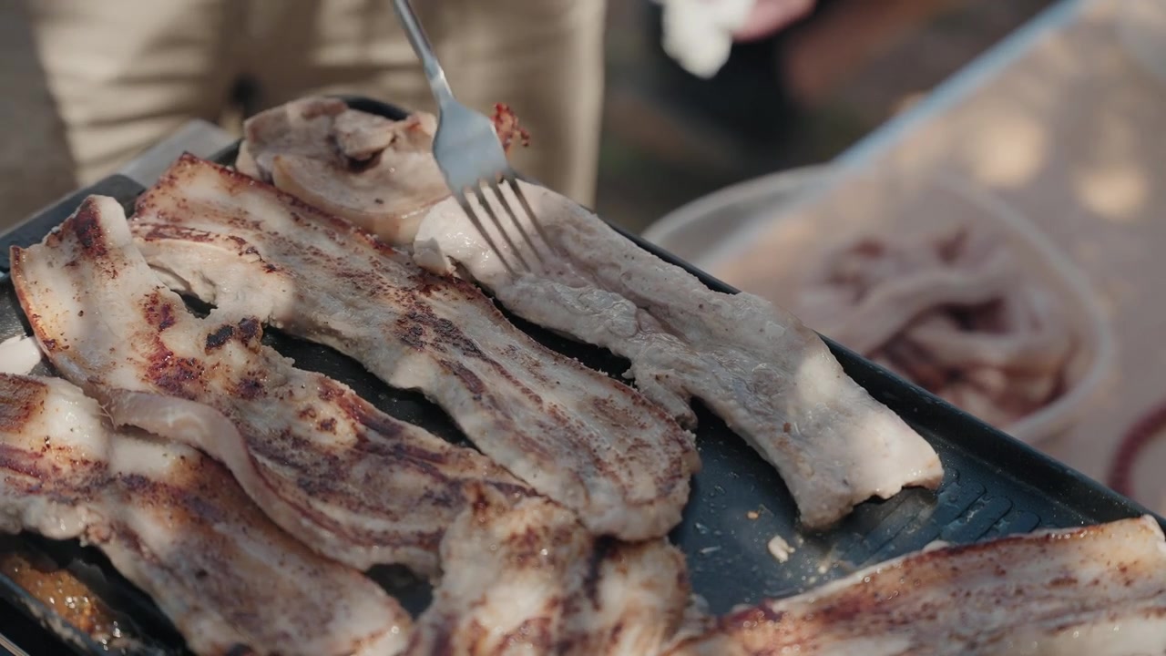 Mano che gira striscioline di pancetta di maiale sfrigolanti su una piastra nera calda con una forchetta.