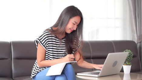 Junger-Asiatischer-Student,-Der-Notizen-Im-Notizbuch-Vom-Laptop-Macht,-Während-Er-Auf-Der-Couch-Sitzt,-Vollbild