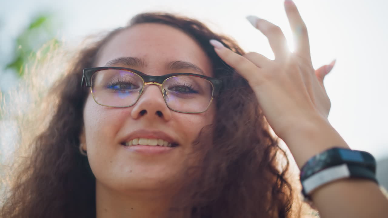 primer plano de una joven con gafas ajustando su cabello mientras posa y sonríe cálidamente frente a la cámara, la luz natural suave crea un brillo suave alrededor de su cara