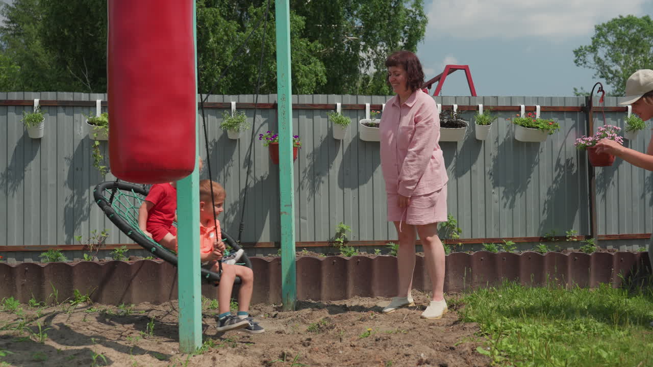 Caucasian Woman With Hat Filming Children On Nest Swing In Backyard, Recording Candid Family Moments With Smartphone, Supportive Adult And Caregiver Nearby, Sunny Garden Setting And Playful Motion