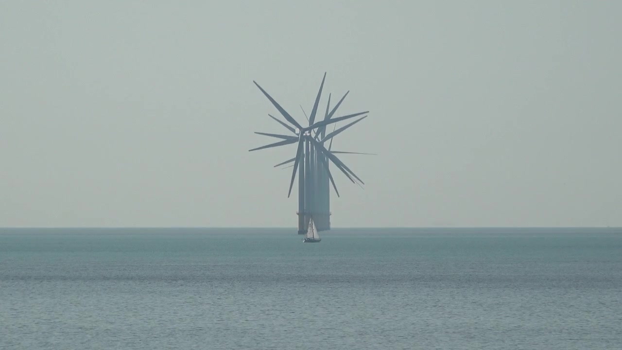 Una piccola barca a vela passa una fila di turbine eoliche che girano al largo su un mare calmo.