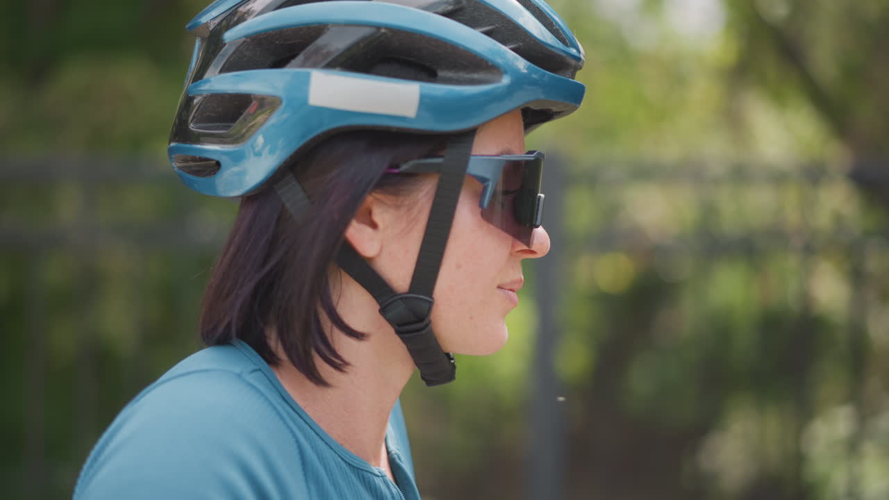 woman wearing helmet and sunglasses in profile, slow urban ride pause, bicycle suggested at frame edge, soft sunlight filtering through trees, blue jersey, helmet strap fastened, calm focused