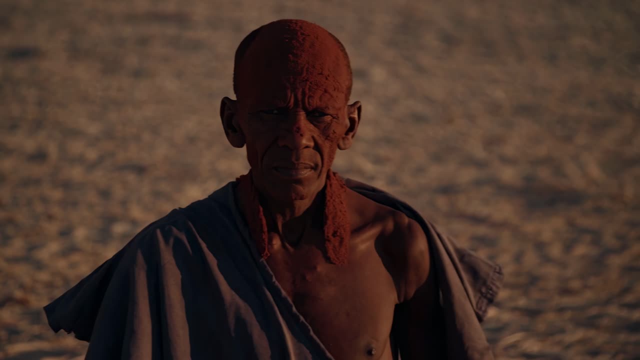 Portrait of an elderly Himba man adorned with traditional ochre on his head and body, wearing a cloth, standing in the Namibian desert at sunset, embodying rich cultural heritage