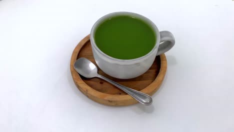 té verde fresco caliente servido en una taza con cuchara sobre una mesa de madera con fondo blanco
