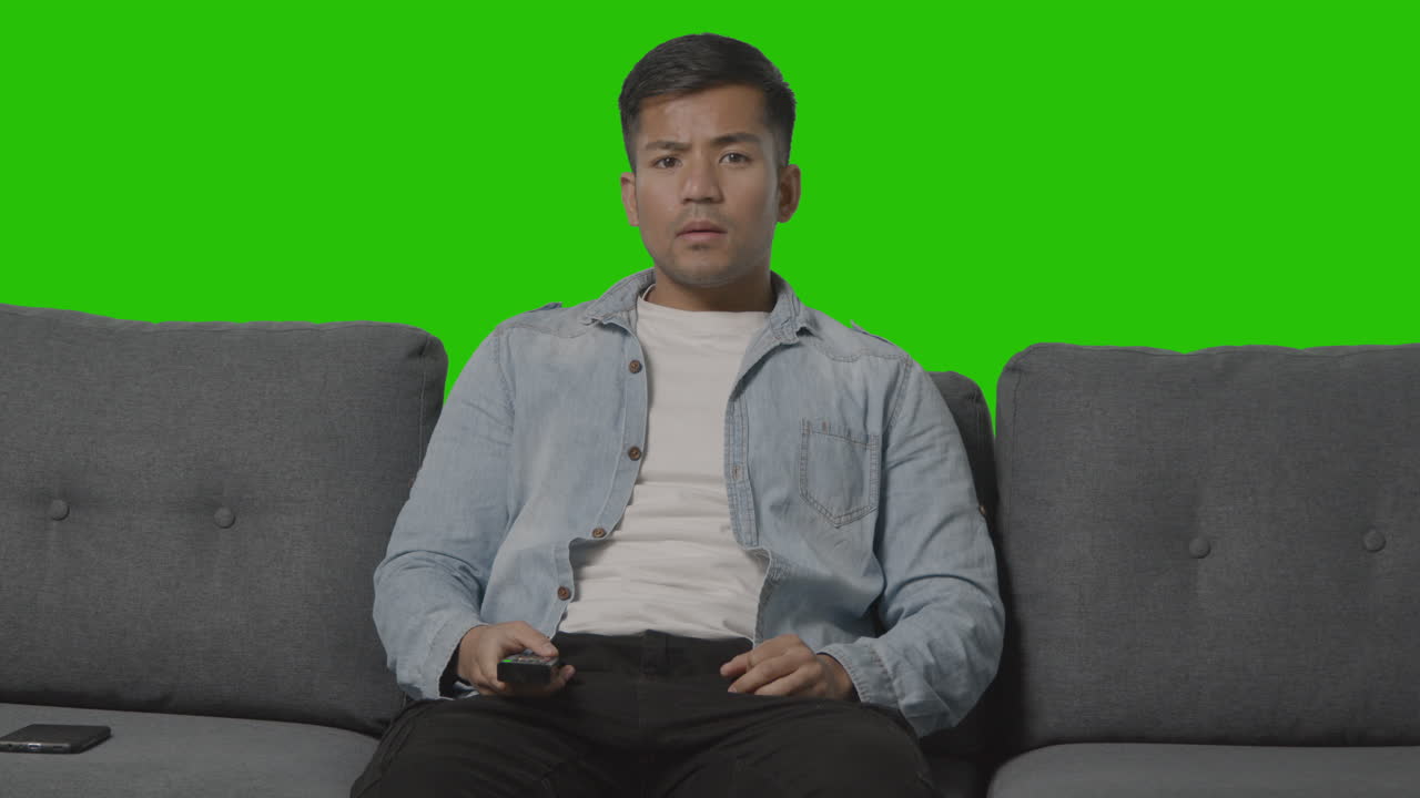 녹색 화면에 대해 tv 채널을 통해 리모콘으로 소파에 앉아 있는 젊은 남자의 스튜디오 샷