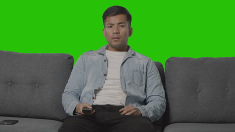 studio shot of young man sitting on sofa with remote control flicking through tv channels against green screen