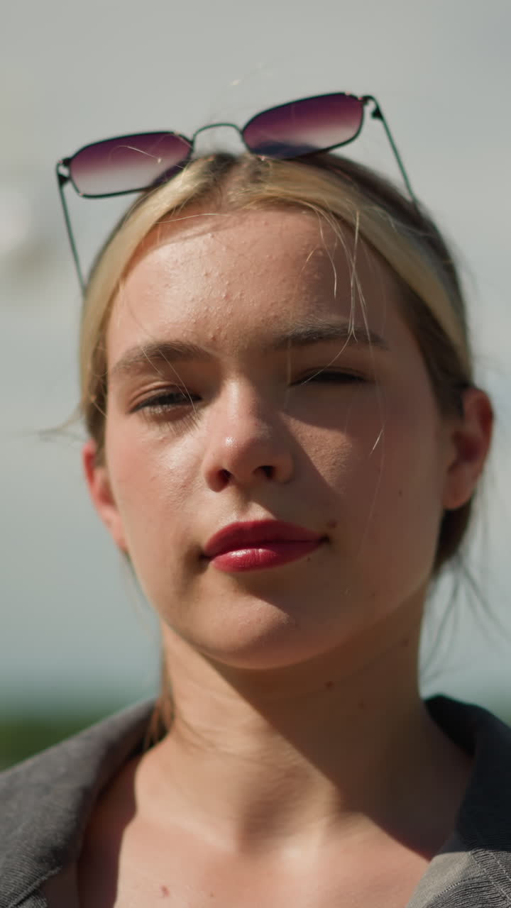 회색 셔츠와 선글라스를 입은 백인 여성은 어깨에 쇼핑 가방을 들고, 약간 뒤로 향하고 집중된 표정으로, 배경은 흐릿하며, 전기 기둥을 특징으로합니다.