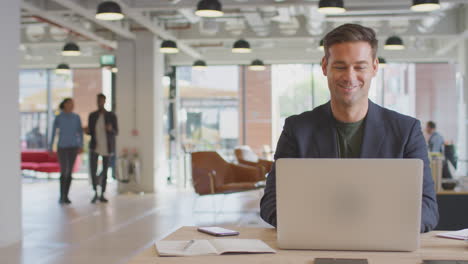 businessman working on laptop at desk with hot drink in modern office with colleagues in background