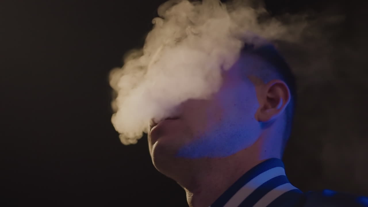 Youthful vapors emerge, Nighttime haze with neon accents, Elegance and attitude in moody vapor scene, Charming young man exhales mist beneath vibrant neon lights with confident pose