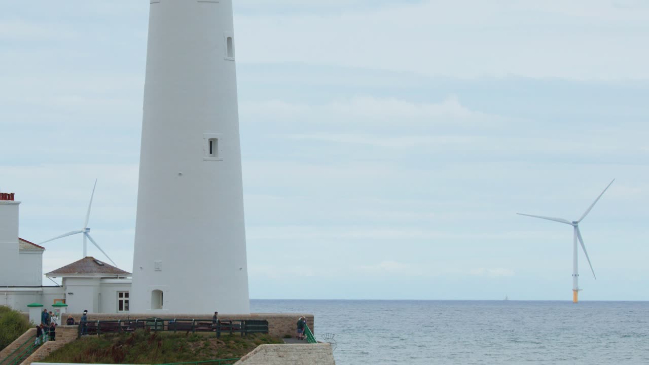 Static wide shot of St. Mary’s Lighthouse and nearby houses with wind turbines in the background under overcast daylight, camera slowly panning right