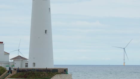 static wide shot of st. mary’s lighthouse and nearby houses with wind turbines in the background under overcast daylight, camera slowly panning right