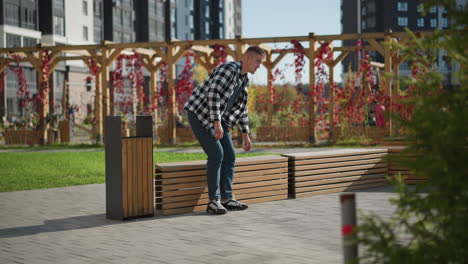 young man in checkered shirt prepares to sit on wooden bench while reaching into front pocket to retrieve medicine, surrounded by decorated wood pergolas and tall modern buildings