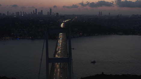 Istanbul-15.-Juli-Märtyrer-Bosporusbrücke-In-Der-Dämmerung-Oder-Nachts-Mit-Der-Silhouette-Der-Stadtsilhouette-Und-Dem-Autoverkehr,-Der-Aus-Den-Lichtern-Der-Stadt-Fließt,-Luftrutsche-Rechts