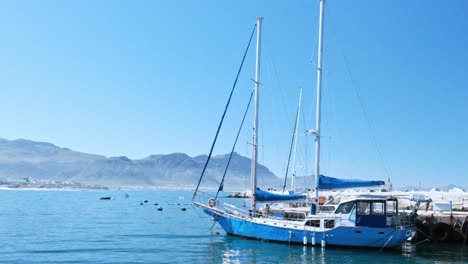 sailing yacht with a blue hull, docked safely in a harbor, on a calm day, with mountains in the distance.