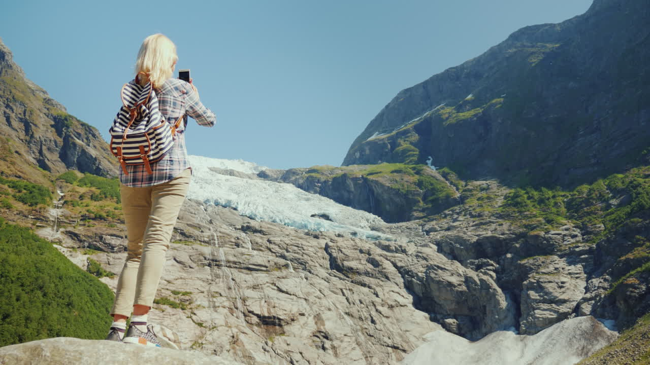 활동적인 여행자 여성이 바위 위에 서서 노르웨이 여행의 아름다운 빙하 사진을 찍습니다.