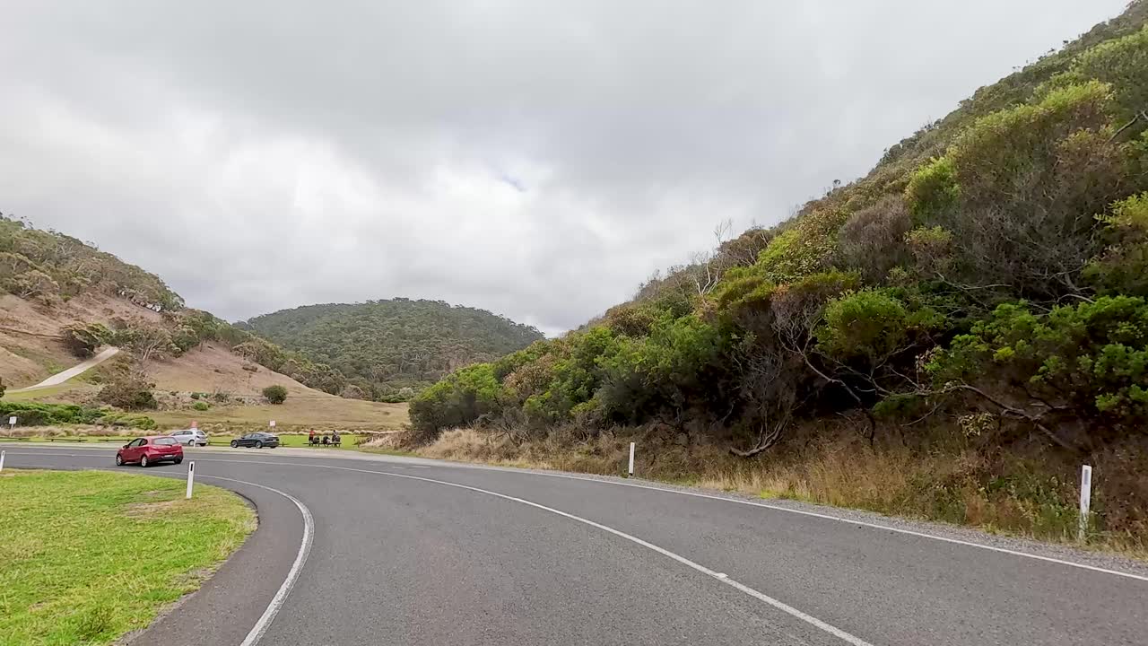 Cars navigate a winding road through lush hills under overcast skies, capturing the serene beauty of Great Ocean Road