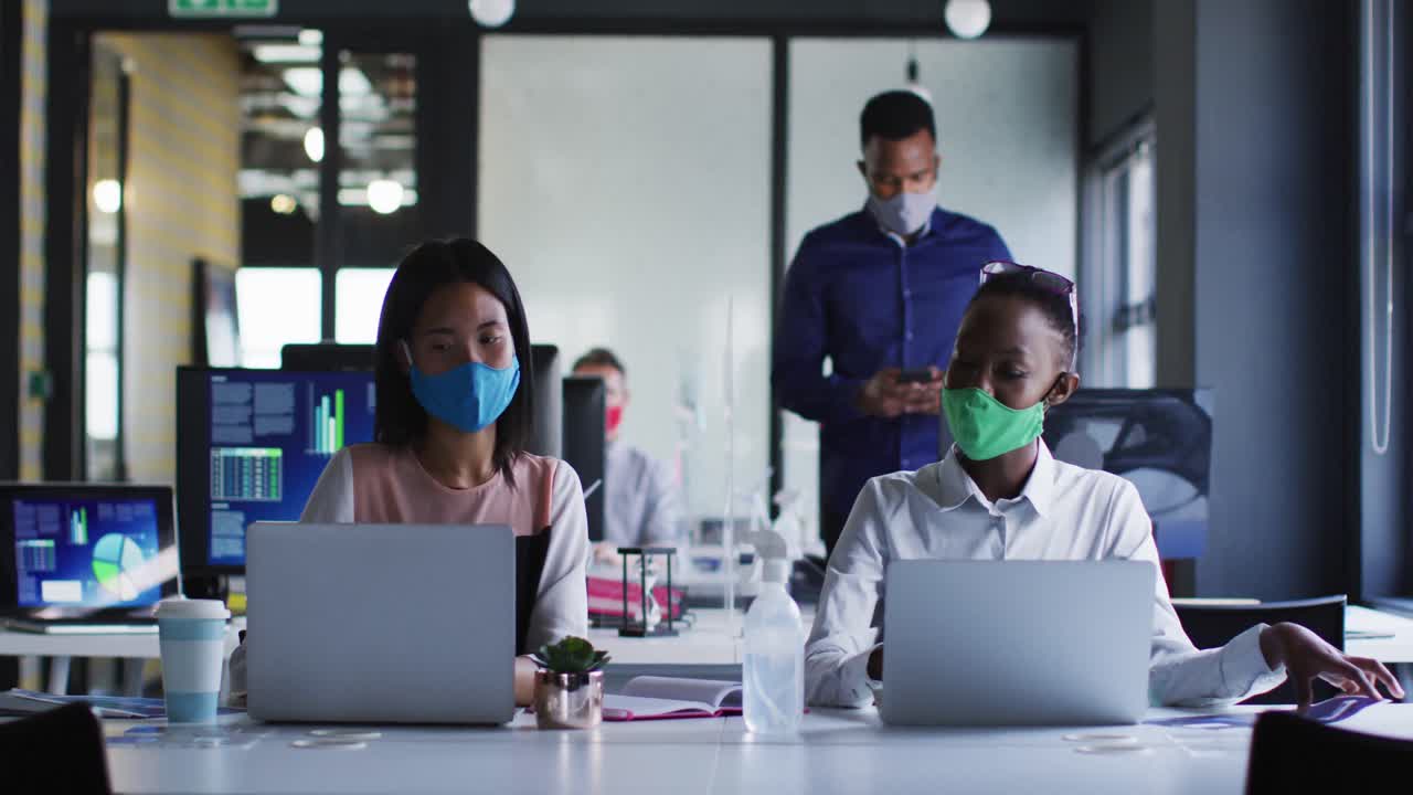 Diverse female colleagues wearing face masks greeting each other by touching elbows at modern office