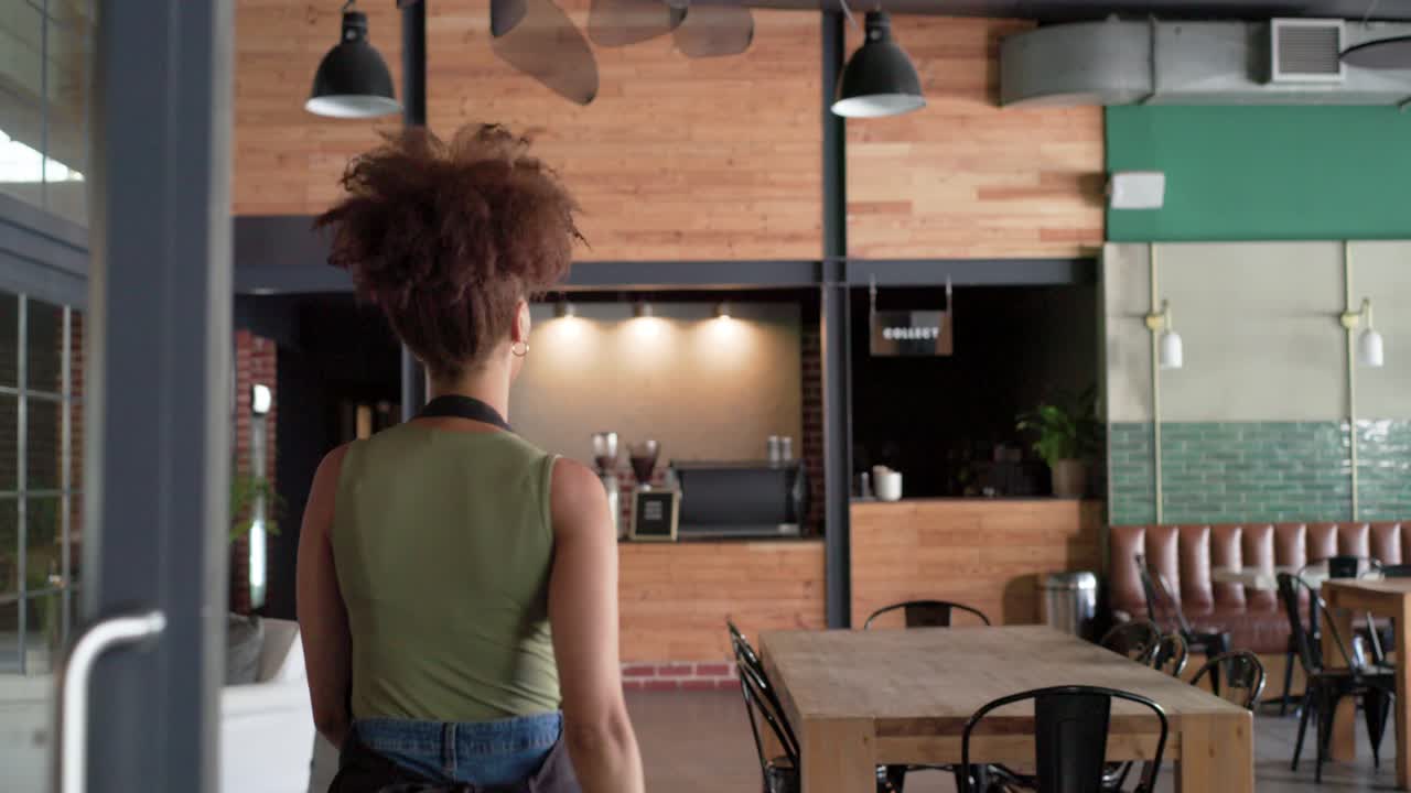 Una barista afroamericana con grembiule gira il cartello chiuso/aperto, apre le porte e prepara le bevande