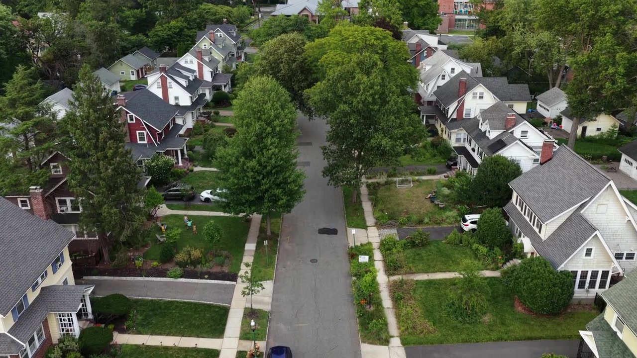 Parking car along tree lined street past large yellow and cream houses in South Orange, New Jersey, manicured lawns and school zone marking line quiet neighborhood street. Aerial drone flyover shot