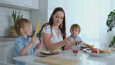 a happy family is a young beautiful mother in a white dress with two sons in blue shirts preparing a white kitchen together slicing vegetables and creating healthy berger for children.