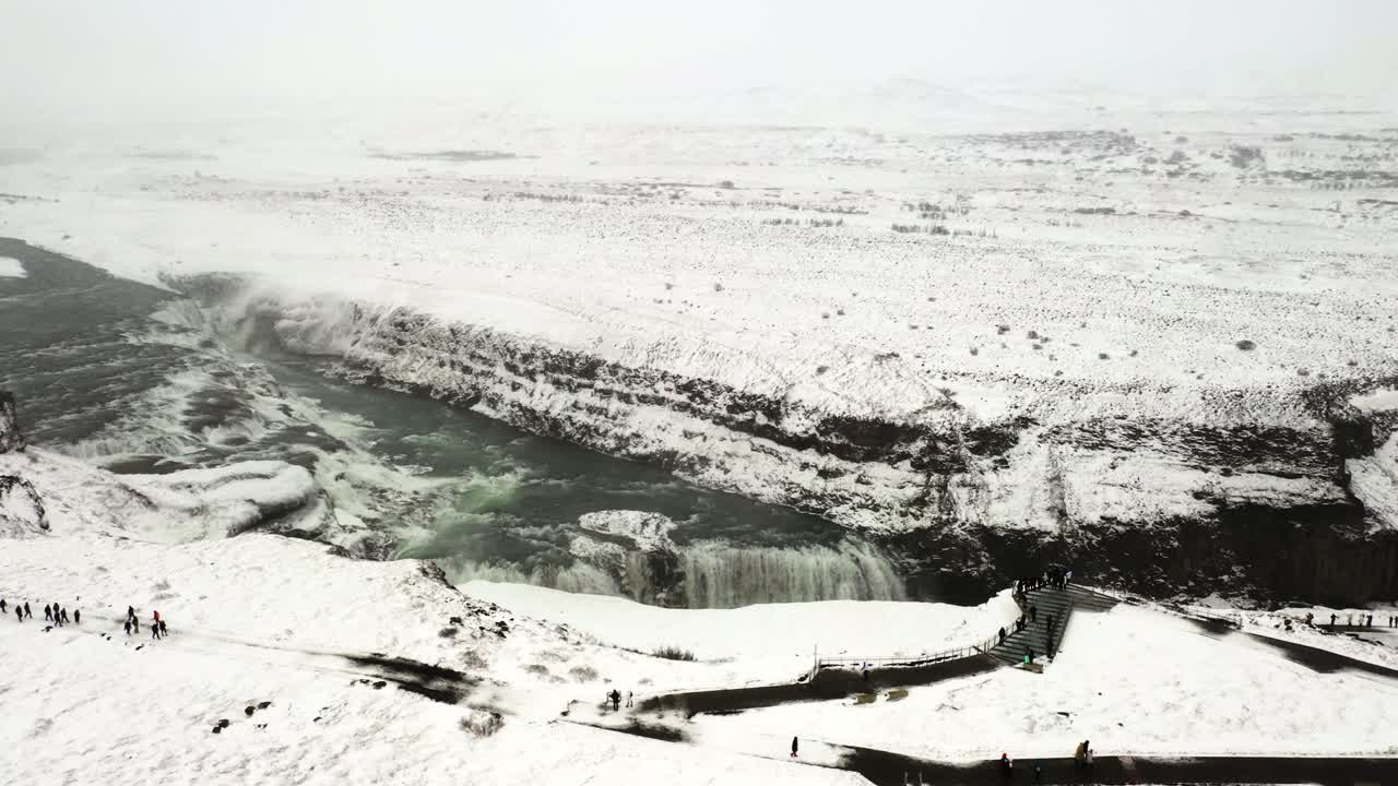 Aerial Footage of Majestic Gullfoss Waterfall, Vast Icy Landscape Surrounded by Snowy Mountain Edges with People Exploring Around