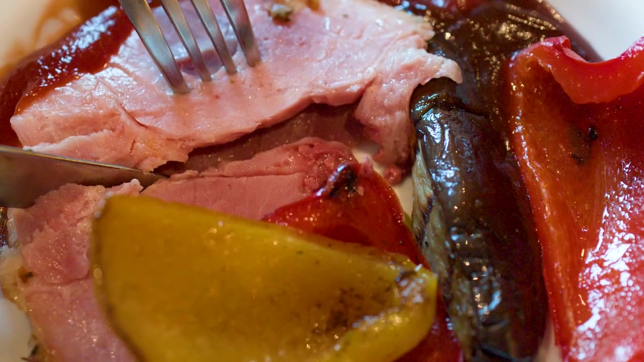Un coltello e una forchetta tagliano del prosciutto arrosto servito con peperoni grigliati e salsa saporita.