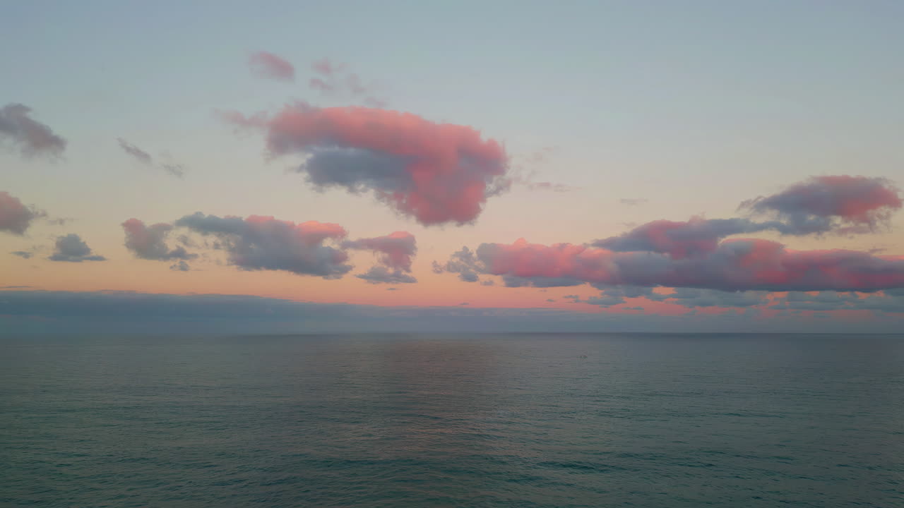 Sunset colors reflect clouds above vast calm ocean in stunning aerial view.