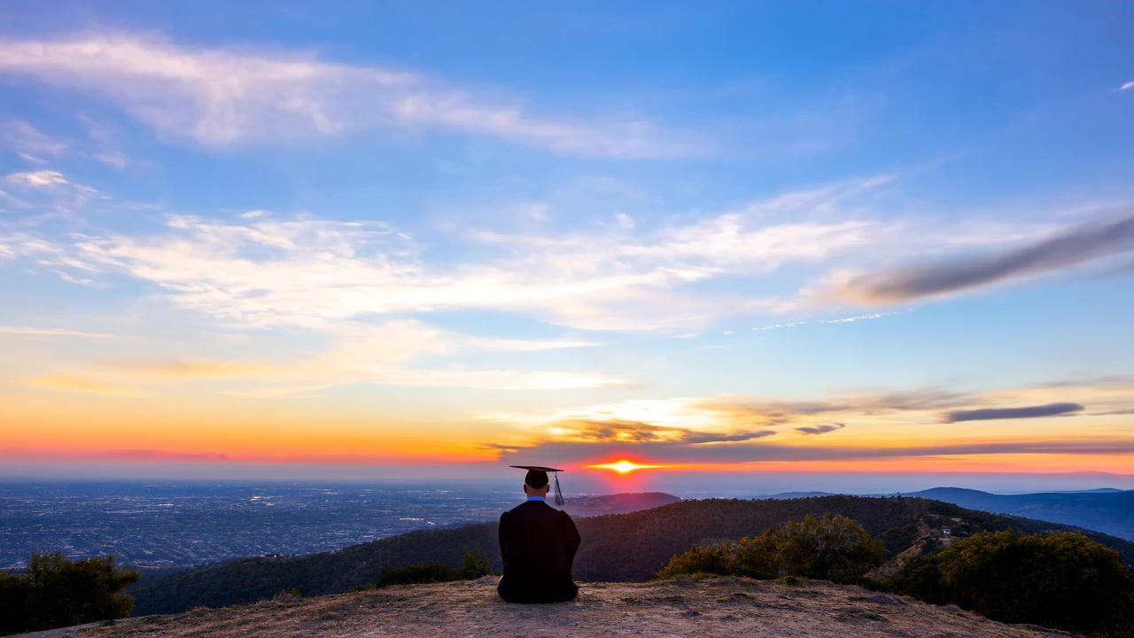 Un graduado vistiendo toga y birrete se sienta en una colina rocosa al amanecer, contemplando un vasto océano y una costa distante mientras sale el sol.