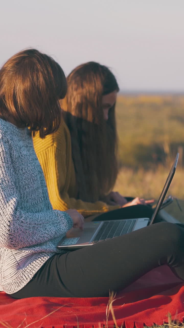 blonde pretty women work on tablet and laptop sitting on red plaid in camp on hilly river bank in autumn evening