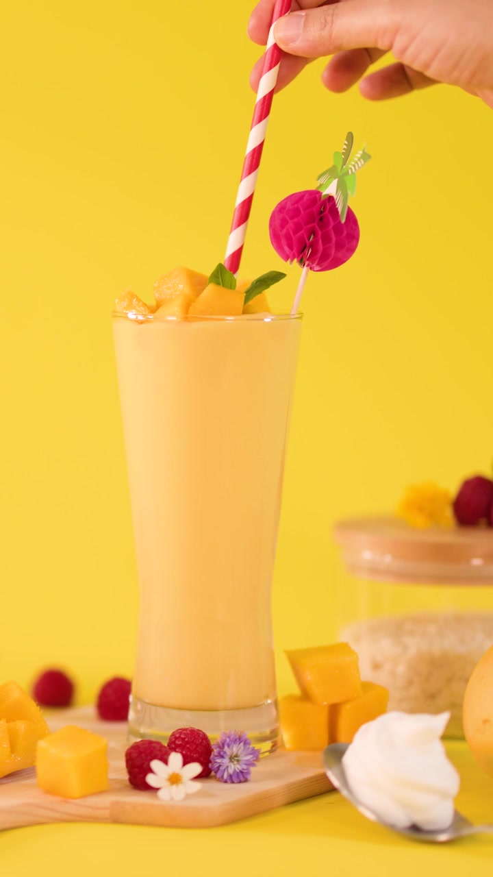 Una mano inserisce una cannuccia a strisce in un alto frullato al mango, circondato da frutta fresca su uno sfondo giallo.