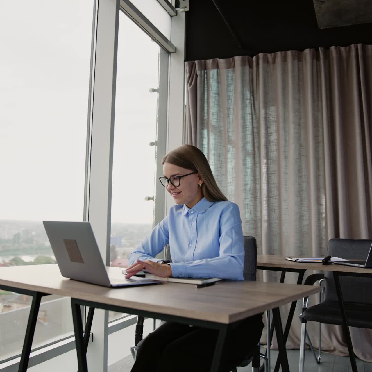 Donna bionda con i capelli lunghi e gli occhiali che lavora al computer seduta alla scrivania. Sfondo luminoso da ufficio con finestre panoramiche