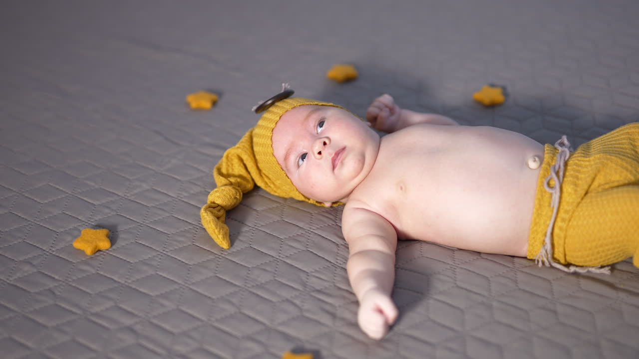 Calm little kid in yellow cap and pants on the bed. Soft toy stars surrounding the baby. Baby boy on the grey backdrop.