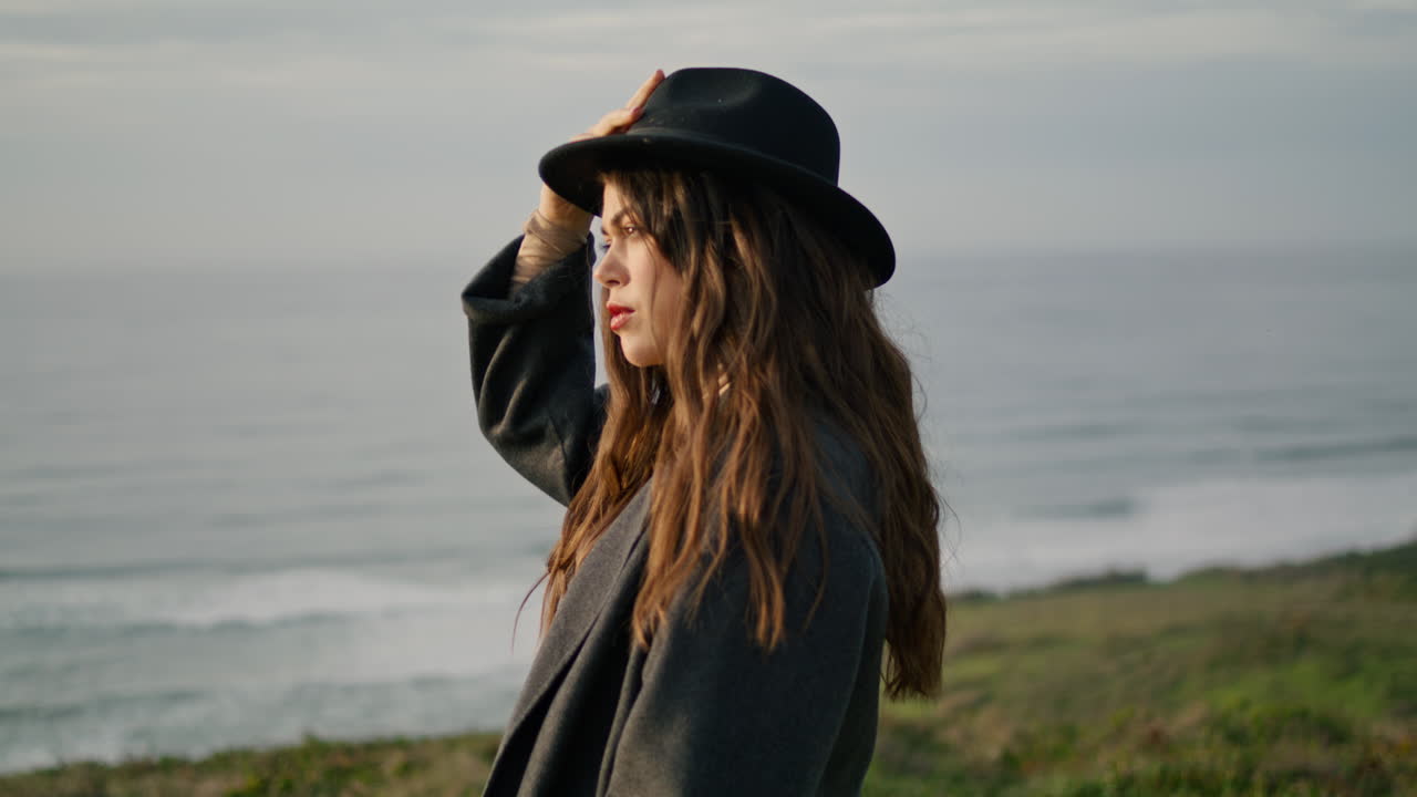 모자를 들고 포즈를 취하는 오션 코스트 어두운 저녁 가까운. 심각한 여성