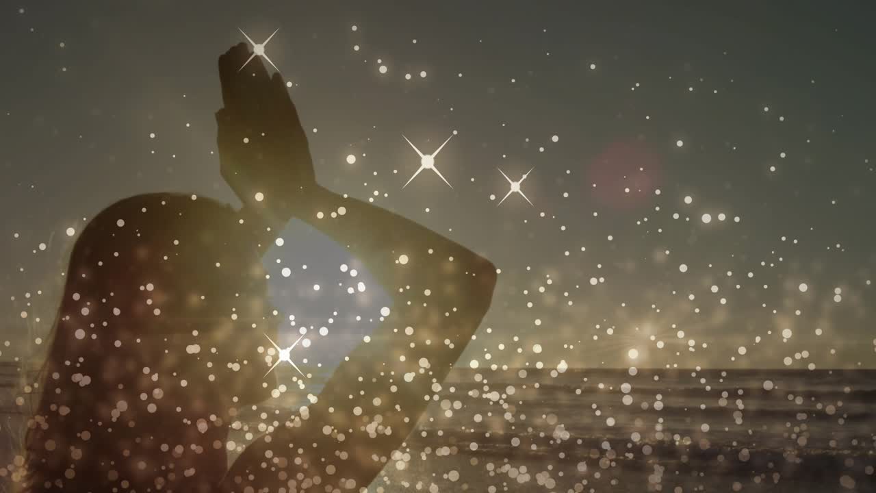Woman holding palms up meditating on shore sun nearing horizon boosting bokeh flares around her