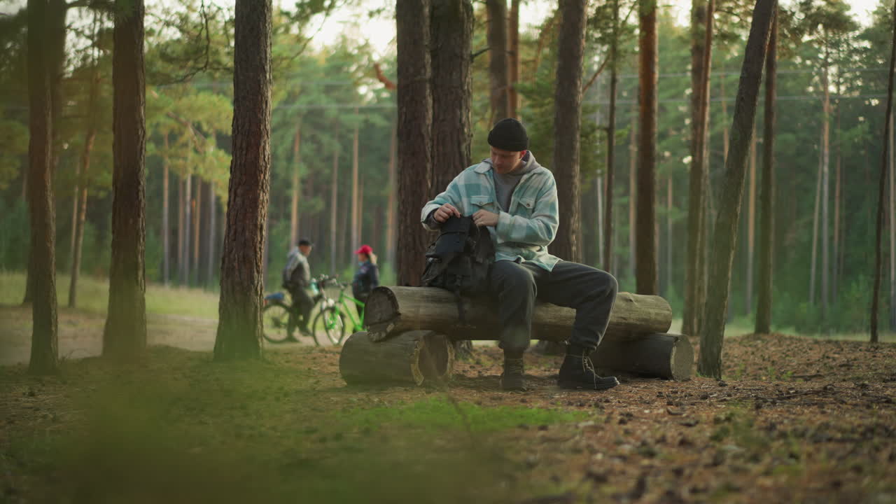 Escursionista caucasico che si riposa su una panchina di legno in una pineta, pausa contemplativa mentre un duo di ciclisti passa, che tira fuori uno snack dallo zaino, una luce dorata e soffusa filtra attraverso gli alti pini, terreno muschioso e silenzio