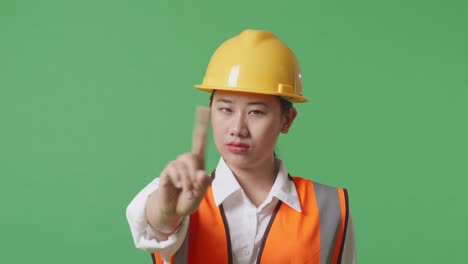 안전 헬을 입은 아시아 여성 엔지니어가 녹색 스크린 배경 스튜디오에 서 있는 동안 지표손가락 표시 없이 불쾌감을 표합니다.