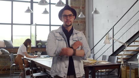 Young-Man-In-A-Hat-And-Jacket-Looks-At-The-Camera-In-A-Tailor-Shop,-In-The-Background-Two-Women-Working-At-A-Table