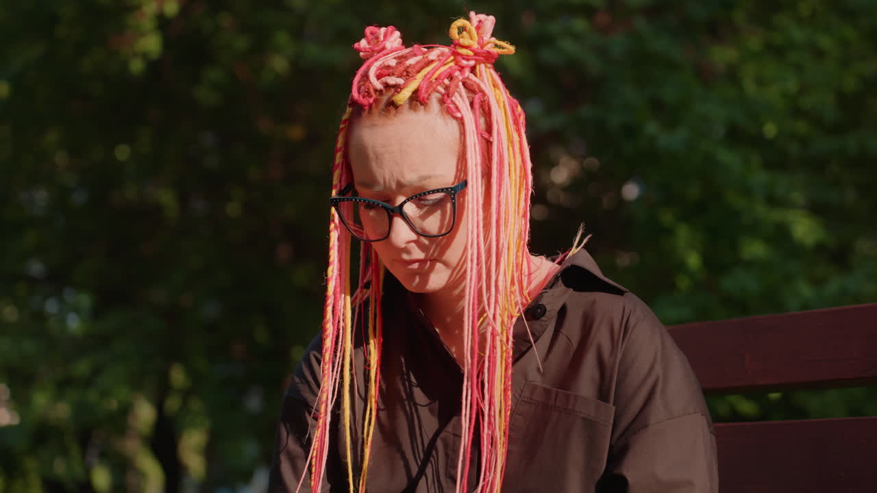 Joven absorta en la lectura de un libro en un banco del parque, lectora con gafas disfrutando de una tarde soleada al aire libre, mujer con el pelo rosa vibrante y gafas sentada a la luz del sol leyendo una novela con tranquilidad