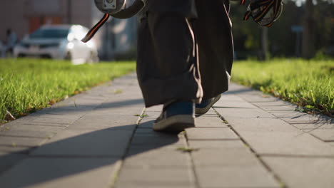 lower half of person walking down paved path wearing dark-colored pants and blue shoe on one foot, low camera angle focusing on textured ground, highlighting casual movement