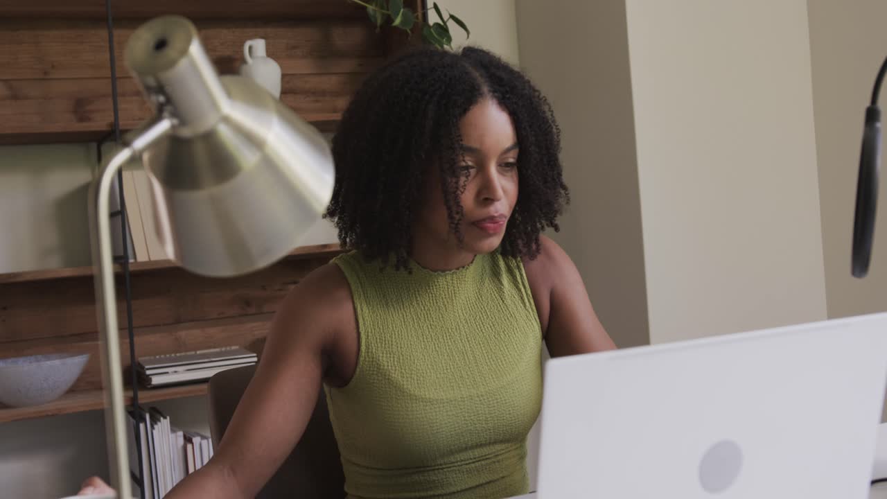 Após um gole, mulher afro-americana adulta colocando a caneca, inclinada e trabalhando em laptop prateado na mesa.