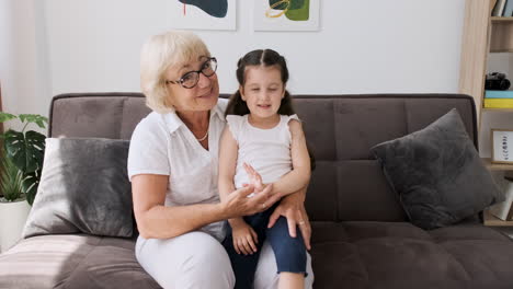 happy grandmother with her pretty little granddaughter sitting on her knees on sofa greeting and talking on video call 1